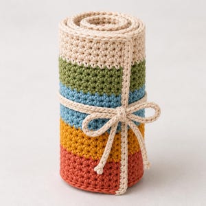 Puede incluir: Un rollo de ganchillo cilíndrico con rayas horizontales en color crema, verde oliva, azul claro, amarillo mostaza y naranja terracota. El rollo está sujeto con un lazo de ganchillo a juego en color crema atado con un nudo en la parte frontal.