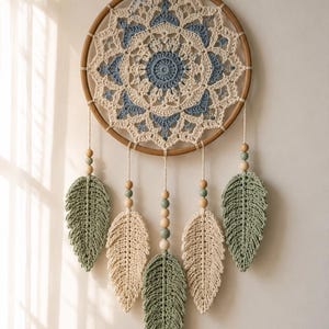 Könnte beinhalten: Ein runder Wandbehang mit einem Holzring und einem gehäkelten Mandala-Muster in Creme und Blau. Fünf gehäkelte blattförmige Anhänger in Creme und Salbeigrün hängen unten, verziert mit kleinen Holzperlen und salbeigrünen Perlen.