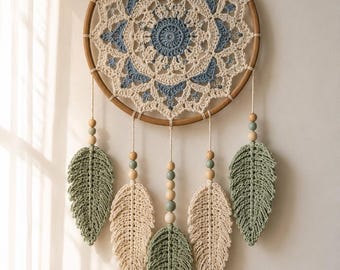 Puede incluir: Un tapiz circular con un aro de madera y un patrón de mandala tejido a ganchillo en crema y azul. Cinco hojas tejidas en crema y verde salvia cuelgan de la parte inferior, decoradas con pequeñas cuentas de madera y verde salvia.