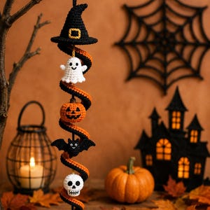 Op de afbeelding: Een hangende gehaakte spiraaldecoratie met een zwarte heksenhoed aan de bovenkant, gevolgd door een wit spookje, een oranje pompoen, een zwarte vleermuis, een witte schedel en een zwarte spin, verbonden door een zwart-oranje spiraal.