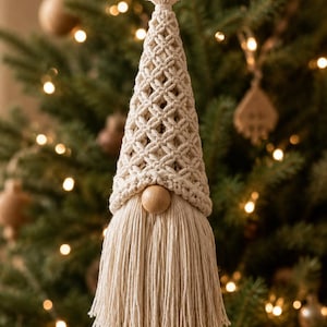 May include: A hanging gnome ornament crafted from off-white macrame cord. The conical hat features a diamond-patterned knot design, while the face includes a smooth, round wooden bead nose above a long, thick, fringed beard made of cotton cord.