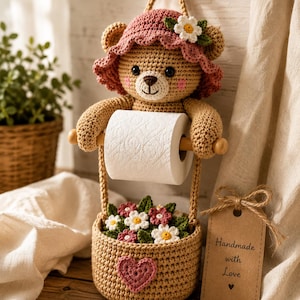May include: A crocheted tan bear wearing a pink hat with a white flower, holding a roll of white toilet paper. Below the bear is a tan crocheted basket filled with small crocheted flowers and a pink heart, with a tag reading Handmade with Love.