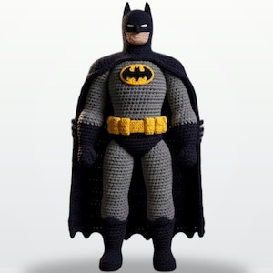 May include: A crocheted doll of a superhero character featuring a dark blue cowl, cape, gloves, and boots. The figure has a grey torso and legs, a yellow utility belt, and a yellow and black oval emblem on the chest. The face has white eye slits.
