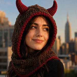 May include: A brown crochet hood featuring two prominent red horns on top. The hood has a thick, textured knit pattern and is finished with a red border along the face opening. The design covers the head and neck, providing a snug fit.