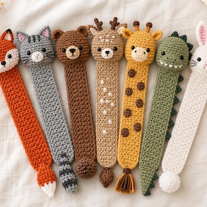 Könnte beinhalten: Eine Sammlung von sieben handgefertigten Häkel-Lesezeichen in Tierform, darunter ein Fuchs, eine graue Katze, ein brauner Bär, ein Reh, eine gelbe Giraffe, ein grüner Dinosaurier und ein weißer Hase. Jedes Lesezeichen hat einen detaillierten Tierkopf.