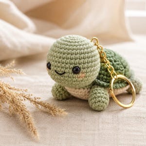 Peut inclure: Un petit porte-clés tortue en crochet avec une tête et un corps vert clair et une carapace vert foncé. La tortue a des yeux en perles noires, un sourire brodé et des joues roses. Un anneau en métal doré est fixé à la carapace.