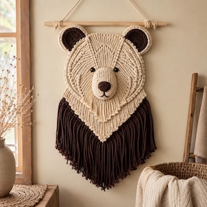 Peut inclure: Une décoration murale en macramé en forme de tête d'ours, réalisée en corde couleur crème avec des accents marron foncé sur les oreilles, le museau et une longue base à franges. La tête d'ours est suspendue à une tige en bois horizontale.