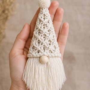 Puede incluir: Un adorno de gnomo de macram&eacute; en color crema que presenta un sombrero triangular con un dise&ntilde;o de nudos en forma de diamante, un peque&ntilde;o pomp&oacute;n en la parte superior, una cuenta de madera lisa como nariz y una larga barba con flecos.