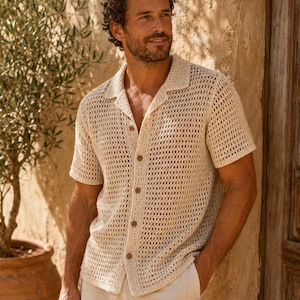May include: A short-sleeved, cream-colored button-down shirt featuring an open-knit crochet texture, a classic collar, and five brown buttons down the front. The garment is constructed with a breathable, grid-like pattern throughout the fabric.