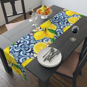 Könnte beinhalten: Tischläufer mit Zitronen- und Blumenmuster. Der Läufer zeigt leuchtend gelbe Zitronen und weiße Blüten vor einem blau-weißen Hintergrund. Der Läufer liegt auf einem dunkelgrauen Holztisch, mit Tellern, Gabeln und Weingläsern.