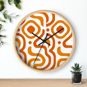 Könnte beinhalten: Eine runde Wanduhr mit hellem Holzrahmen. Das Zifferblatt hat einen weißen Hintergrund mit einem sich wiederholenden Muster aus orangefarbenen und gelben abstrakten Formen. Schwarze Zeiger zeigen die Uhrzeit an.