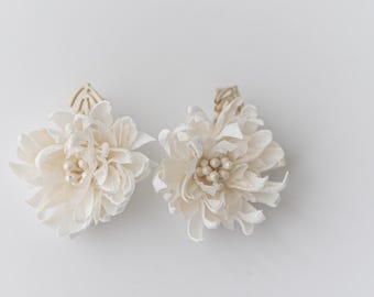 以下が含まれることがあります： パールがあしらわれた白い布の花のヘアクリップ2個。クリップは金色の金属の茎に取り付けられています。