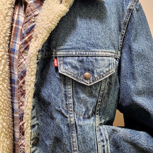 May include: A blue denim jacket with a cream-colored sherpa lining, featuring a chest pocket with a button closure and a small red Levi's brand tag. The jacket is worn over a plaid button-down shirt with blue, brown, and white colors.