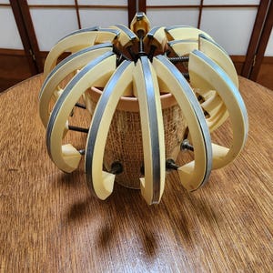 May include: An antique plant pot holder with a cream-colored, curved design and silver metal accents. The holder is open and adjustable, with a woven basket-style pot inside. The holder is sitting on a wooden table.