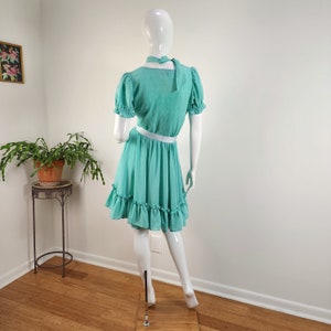 Puede incluir: Un vestido azul claro con una cinta blanca alrededor de la cintura. El vestido tiene un dobladillo con volantes y mangas cortas abullonadas. El vestido est&aacute; en un maniqu&iacute; blanco.