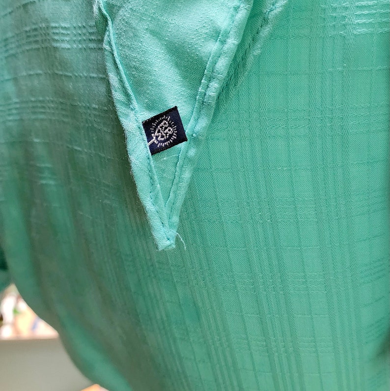 Puede incluir: Una tela de algod&oacute;n verde claro con una peque&ntilde;a etiqueta negra con un logotipo. La tela tiene un patr&oacute;n de cuadr&iacute;cula.