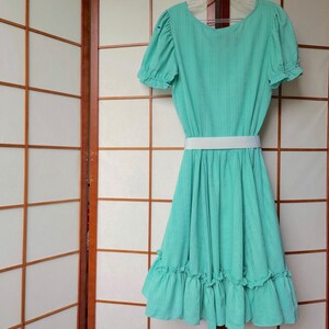 Puede incluir: Un vestido verde menta con un cintur&oacute;n blanco y un dobladillo con volantes. El vestido tiene mangas cortas y un escote redondo.