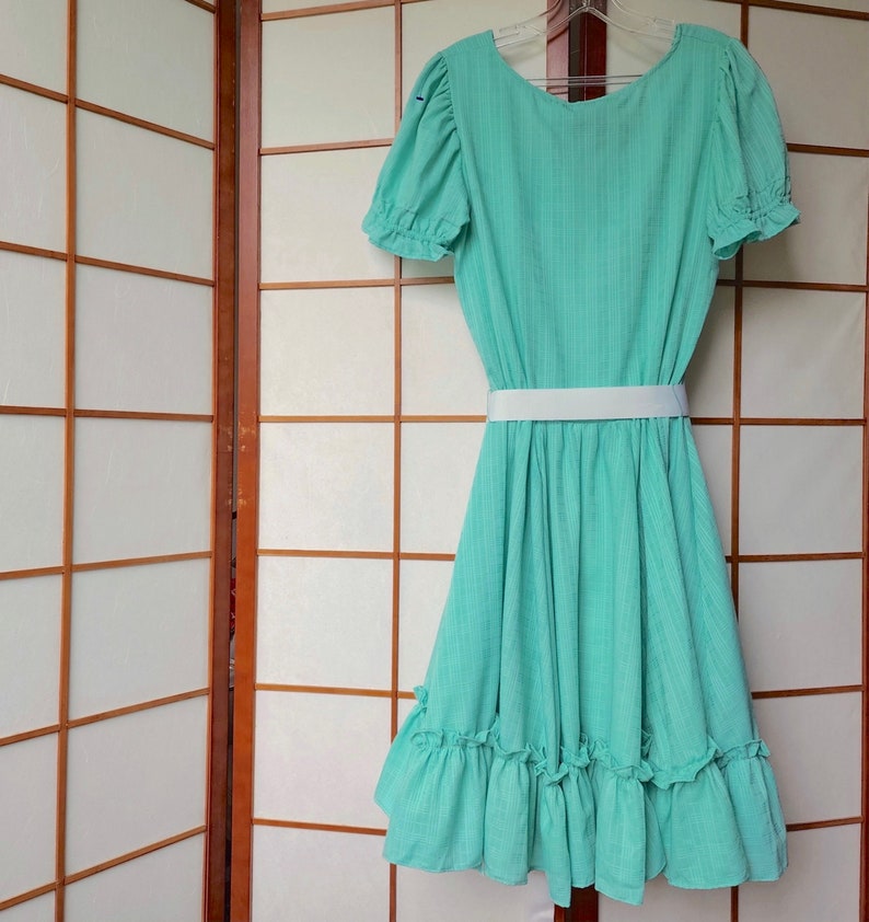 Puede incluir: Un vestido verde menta con un cintur&oacute;n blanco y un dobladillo con volantes. El vestido tiene mangas cortas y un escote redondo.
