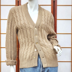 May include: A beige cable-knit cardigan with a V-neck and button fastening. The long sleeves have turned-up cuffs. The sweater is displayed on a mannequin. The sweater is a neutral colour.
