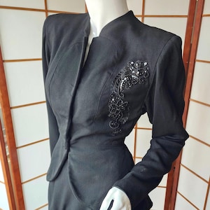 May include: A black long-sleeved jacket featuring a mandarin-style collar and a single front button closure. The left side of the chest is adorned with an intricate black lace appliqué embellished with matching black sequins and beads.