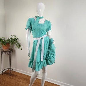 Puede incluir: Un vestido azul claro con una faja blanca y un lazo blanco en el cuello. El vestido tiene un dobladillo con volantes y un corpi&ntilde;o ajustado.