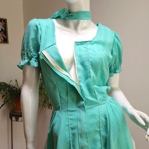 Puede incluir: Un vestido azul turquesa con una cremallera en la parte delantera y mangas cortas abullonadas. El vestido tiene un escote cuadrado y un corpi&ntilde;o ajustado.