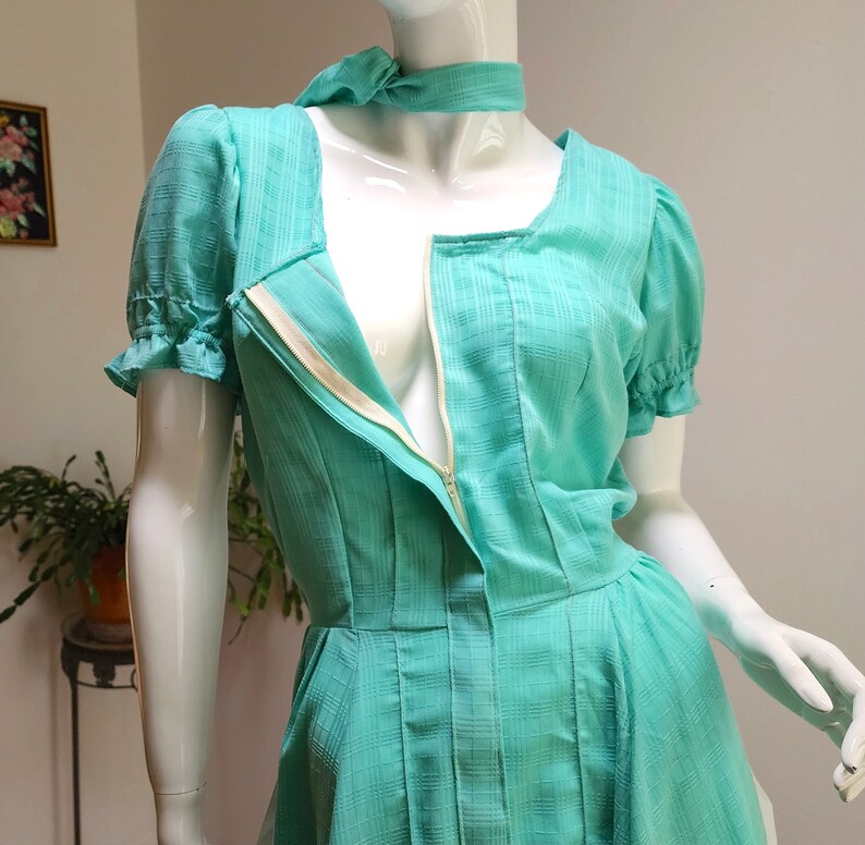 Puede incluir: Un vestido azul turquesa con una cremallera en la parte delantera y mangas cortas abullonadas. El vestido tiene un escote cuadrado y un corpi&ntilde;o ajustado.