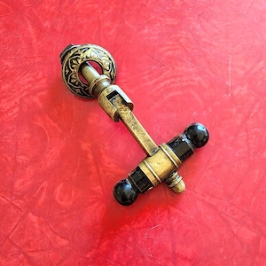 May include: An antique-style cabinet pull with a gold finish and black accents. The pull features a decorative circular backplate with an ornate design, a rectangular stem, and a handle with two black spherical knobs. The hardware is set against a red background.