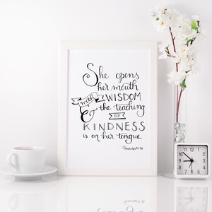 May include: A white framed print with a black and white hand-lettered quote: "She opens her mouth with wisdom & the teaching of kindness is on her tongue." - Proverbs 31:26
