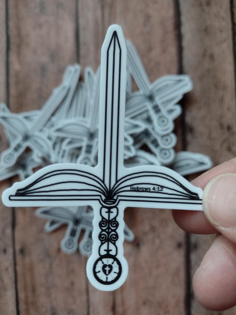 Peut inclure: Autocollant blanc en forme d'&eacute;p&eacute;e avec une garde en forme de croix et un motif de livre ouvert. Le texte "H&eacute;breux 4:12" est visible sur l'autocollant. L'autocollant est tenu par les doigts d'une personne.
