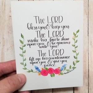 May include: A white card with a floral border and a handwritten quote from the Bible, Numbers 6:24-26. The quote reads: "The LORD bless you and keep you. The LORD make his face to shine upon you and be gracious unto you. The LORD lift up his countenance upon you and give you peace."
