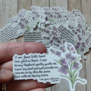 May include: A collection of stickers featuring a floral design with purple flowers and green leaves. The stickers have the text "I am Jesus little lamb" and other religious text. The stickers are on a wooden surface.