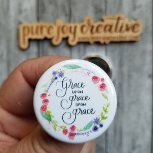 May include: White button with a floral wreath border and the text "Grace upon grace upon grace" in black calligraphy.