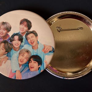 May include: A round button pin featuring a group of smiling people in various pastel-colored shirts. The pin has a glossy finish and a metal pin back. The background is a light beige color.