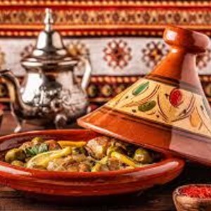 Puede incluir: Un plato tradicional marroqu&iacute; de tagine con tapa c&oacute;nica, con una vibrante combinaci&oacute;n de colores naranja-marr&oacute;n. El plato est&aacute; lleno de un guiso, y en el fondo se ve una tetera plateada. La escena est&aacute; ambientada sobre una superficie de madera.