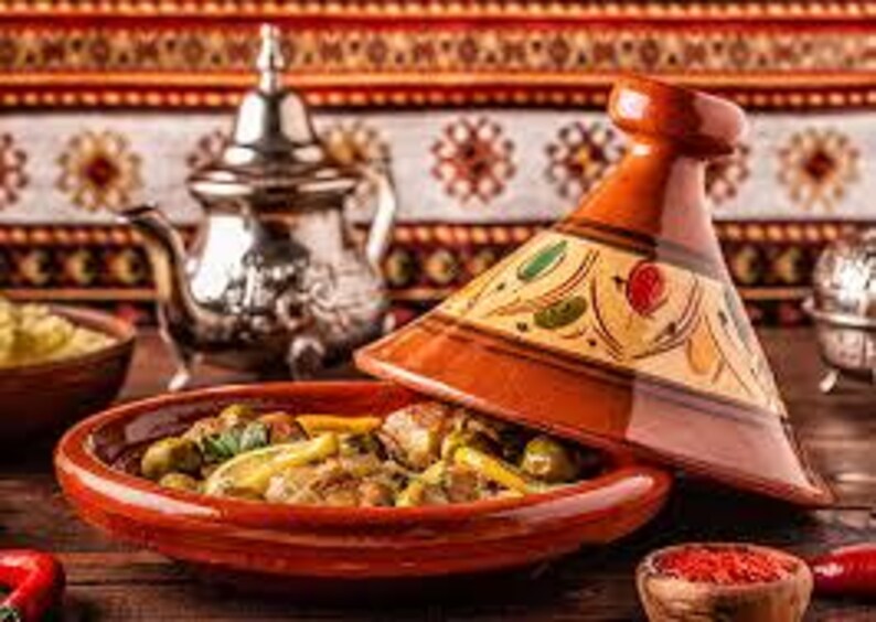 Puede incluir: Un plato tradicional marroqu&iacute; de tagine con tapa c&oacute;nica, con una vibrante combinaci&oacute;n de colores naranja-marr&oacute;n. El plato est&aacute; lleno de un guiso, y en el fondo se ve una tetera plateada. La escena est&aacute; ambientada sobre una superficie de madera.