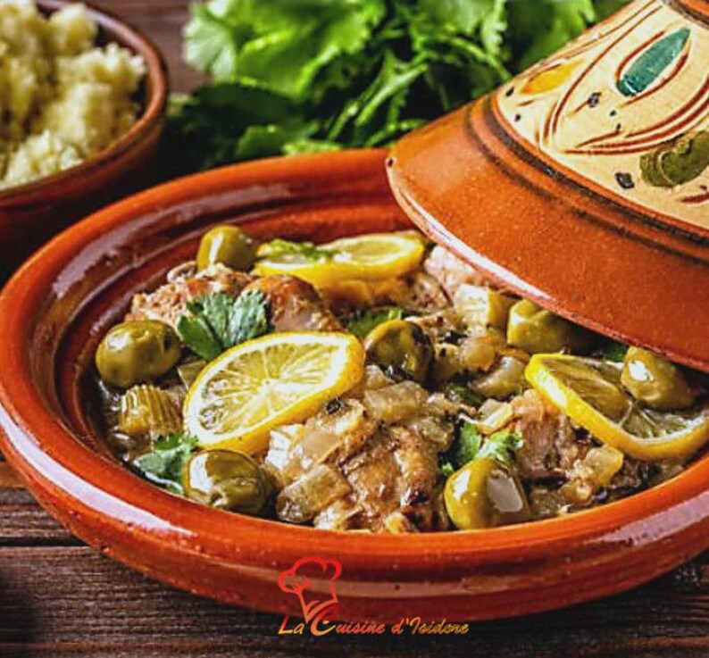 Puede incluir: Un plato de tagine marroqu&iacute; con aceitunas, rodajas de lim&oacute;n y hierbas, servido en una olla de terracota con tapa decorativa. El plato se presenta sobre una superficie de madera, con un cuenco de cusc&uacute;s al fondo. Incluye el texto "La Cuisine d'Isidore".