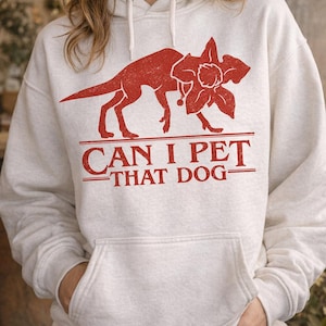 May include: A light gray hoodie with a red graphic of a dog-like creature with a flower. The text "CAN I PET THAT DOG" is printed below the graphic in red. The hoodie has a front pocket and a drawstring hood.