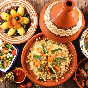 Puede incluir: Vista a&eacute;rea de una variedad de platos, incluyendo cusc&uacute;s, ensaladas y guisos, dispuestos sobre una mesa de madera r&uacute;stica. Los platos presentan verduras vibrantes, aceitunas y albaricoques. Tambi&eacute;n hay una olla de tagine tradicional.