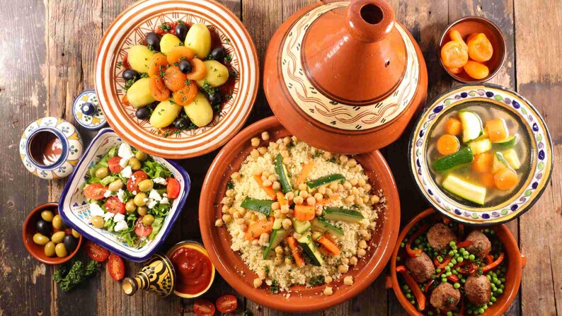 Puede incluir: Vista a&eacute;rea de una variedad de platos, incluyendo cusc&uacute;s, ensaladas y guisos, dispuestos sobre una mesa de madera r&uacute;stica. Los platos presentan verduras vibrantes, aceitunas y albaricoques. Tambi&eacute;n hay una olla de tagine tradicional.