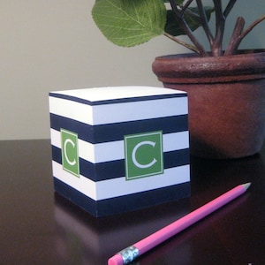 May include: A black and white striped notepad with a green letter "C" on the front. The notepad is sitting on a wooden table next to a potted plant.