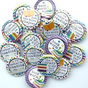 May include: A collection of round, colorful buttons with book-themed designs. Each button features text like "I'd rather be reading," "Book Worm," and "Just one more chapter." The buttons have illustrations of books, hearts, and a green worm.