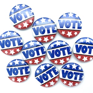 May include: A collection of round buttons with a patriotic design. Each button features the word "VOTE" in white letters against a blue background, surrounded by white stars and red stripes. The buttons are scattered on a white surface.