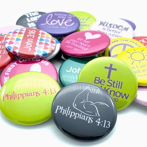 May include: A collection of colorful round buttons with inspirational quotes and bible verses. The buttons feature various colors, including red, pink, yellow, green, and purple. Some of the quotes include "John 3:16", "Be Still and Know", "Wisdom is better than rubies", and "Let not your heart be troubled".
