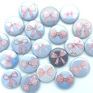 May include: A collection of round, decorative buttons featuring pink bows on a light blue background. The bows have various patterns, including polka dots, stripes, and checkered designs. The buttons are arranged on a white surface.
