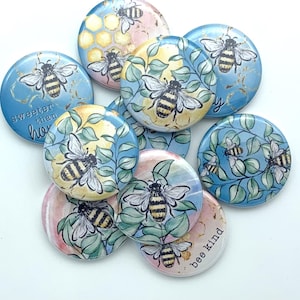 May include: A set of nine round, blue and pink buttons with bee and honeycomb designs. The buttons feature watercolor illustrations of bees, honeycombs, and leaves. Some buttons have text that reads "sweeter than honey" and "bee kind".