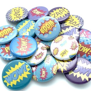May include: A collection of colorful round buttons with various comic book sound effects like "POW!" and "BOOM!" printed on them. The buttons are blue, yellow, red, white, and purple.