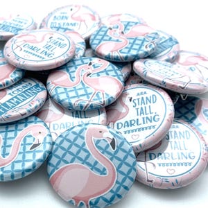 May include: A collection of pink flamingo themed button pins with various sayings, including "Stand Tall, Darling" and "Make Today Flaming".