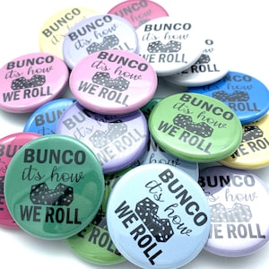 May include: A collection of colorful round buttons with the text "BUNCO it's how WE ROLL" in black. The buttons are in various pastel colors, including pink, blue, green, and yellow. The image shows a close-up of the buttons.