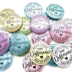 May include: A collection of colorful round buttons with the text "Never Forget The Difference You've Made" and a small heart in the center. The buttons are in various pastel colors, including pink, blue, yellow, and green.